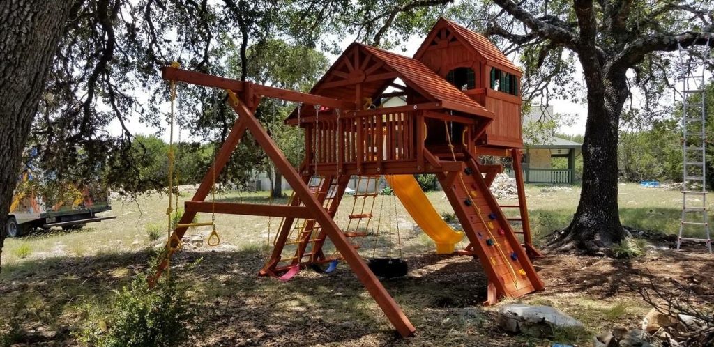 Two-tower playset with swings and a climbing wall installed under live oaks