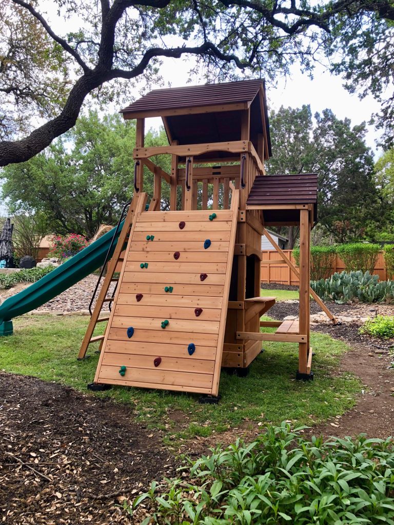 Tall cedar playset with a rock-climbing wall installed in a landscaped backyard