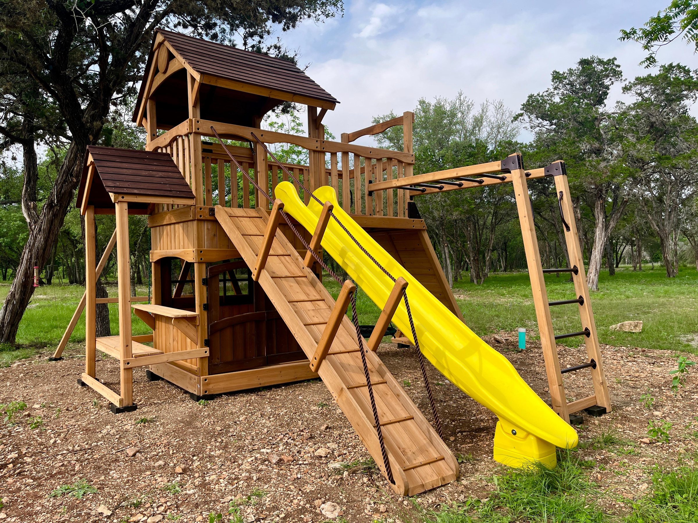 Cedar playset with a yellow slide, climbing ramp, and monkey bars installed among Hill Country oaks